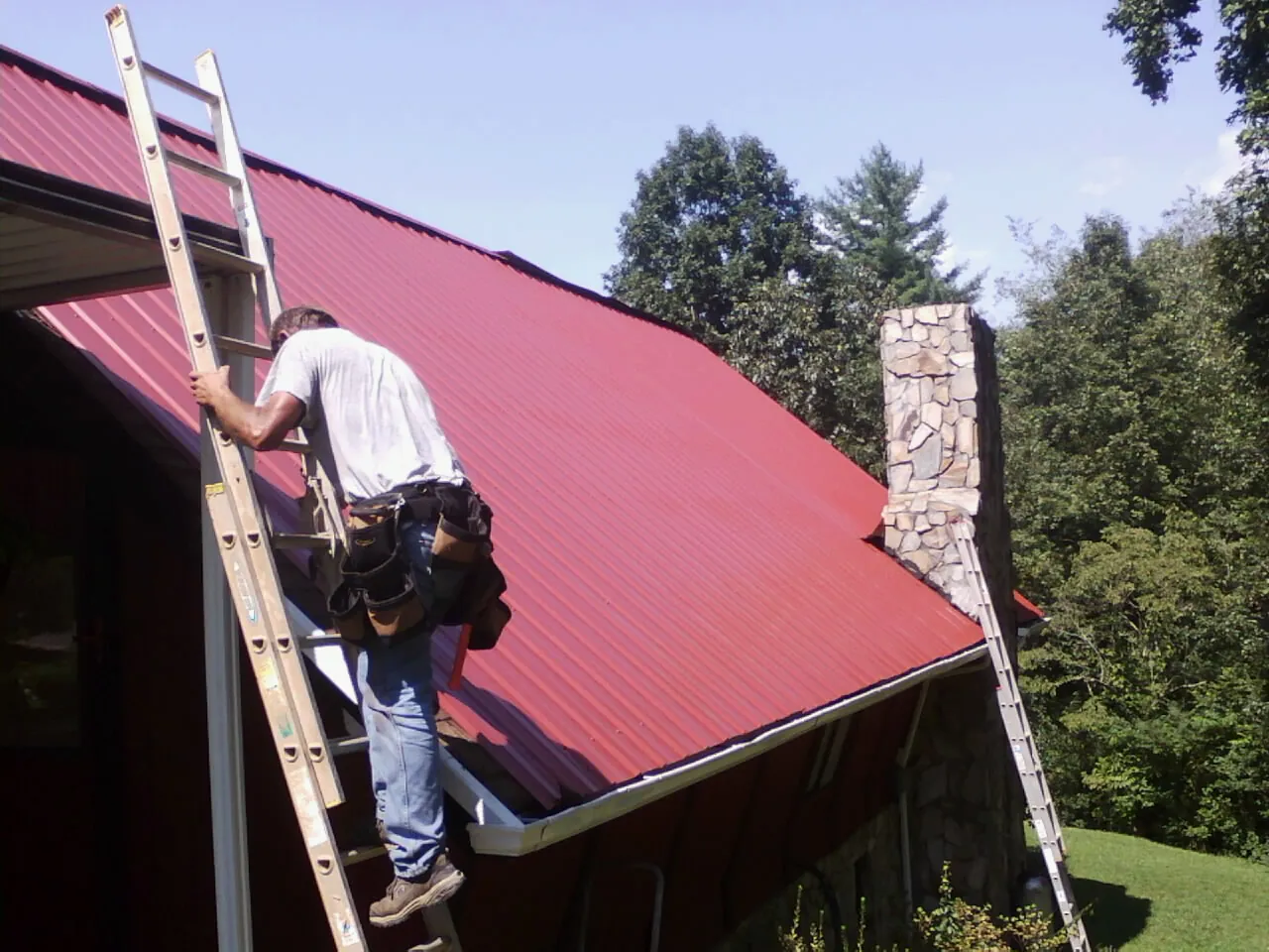 Roofer on a ladder beside a red metal roof and stone chimney — Soffit & Fascia Repair by Roofing Brothers in Boyertown